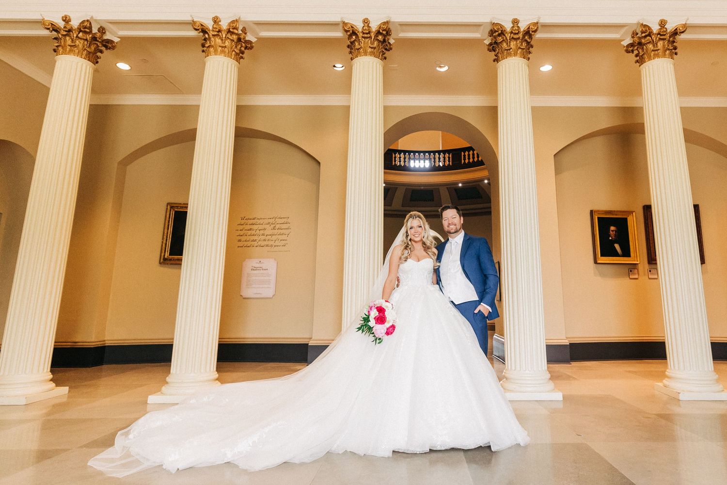A bride and groom pose elegantly between tall white columns in an ornate indoor venue.