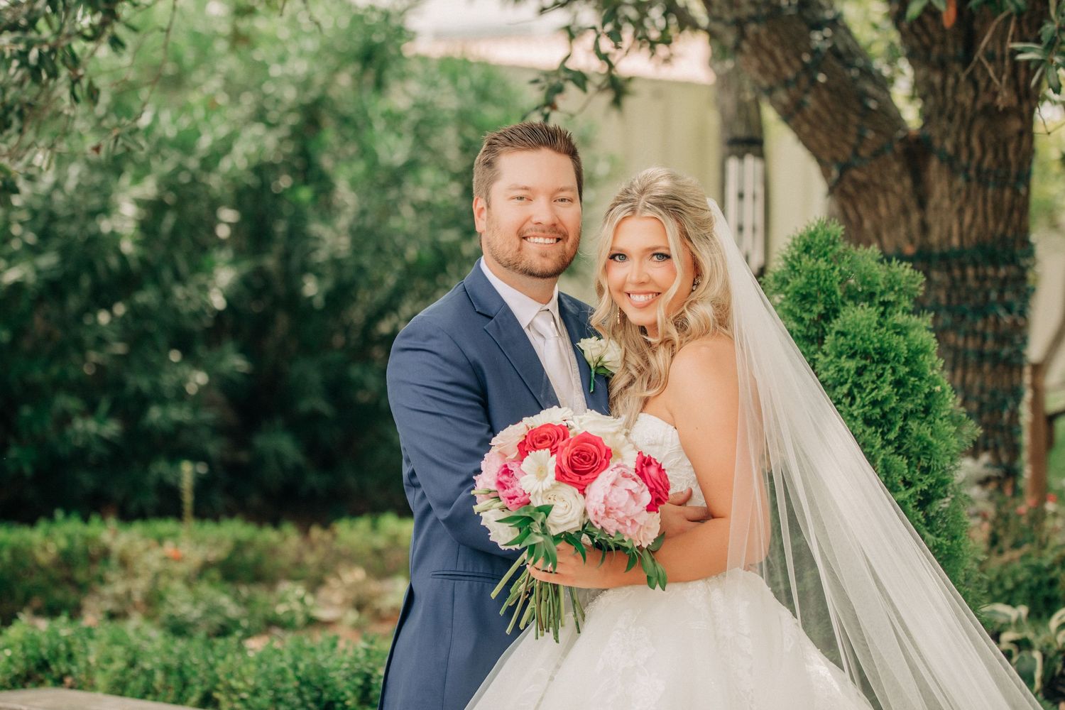 A bride in a white gown holds a pink floral bouquet while standing with her groom in a navy suit in a garden setting.