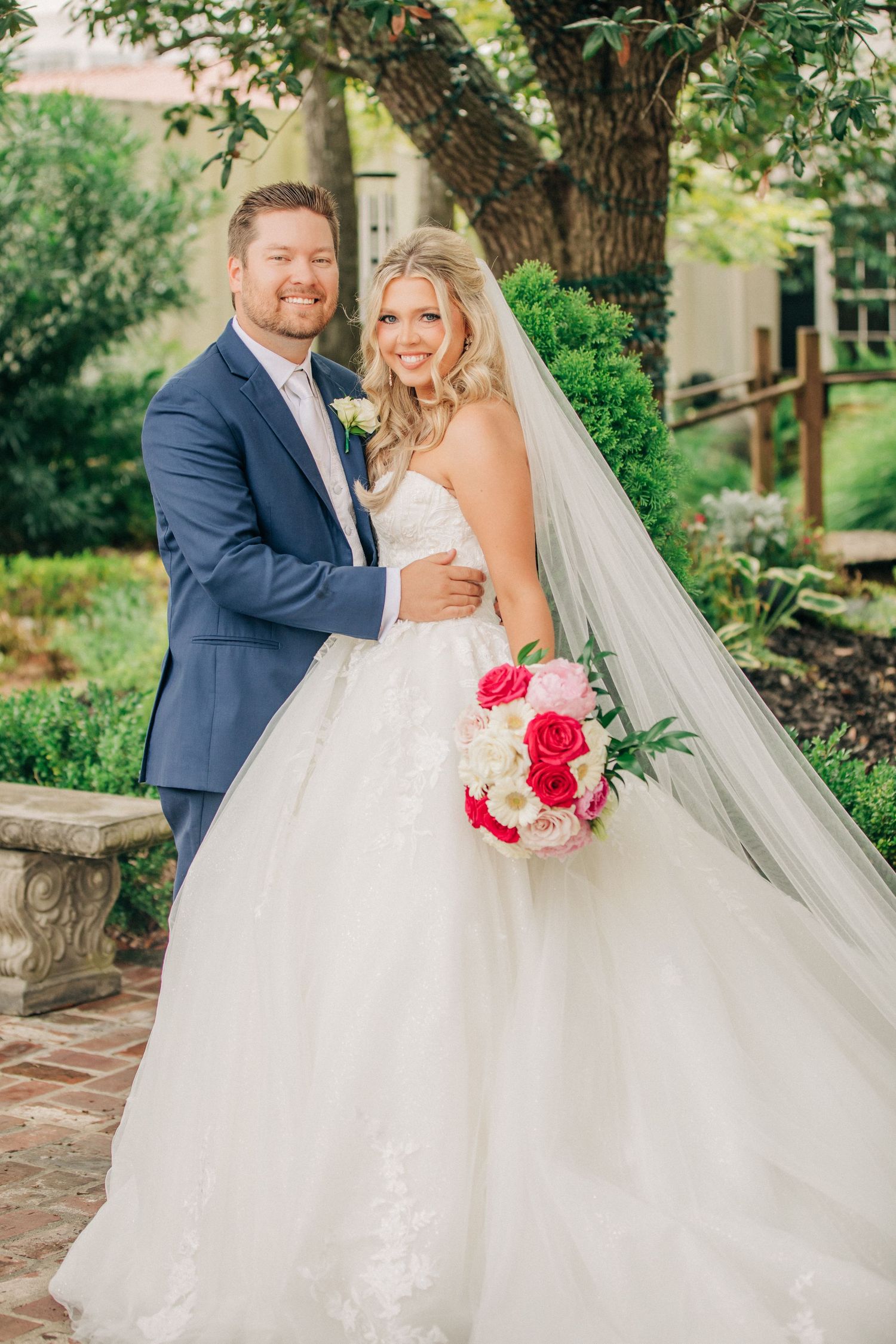 A couple poses for a wedding photo in a garden with a beautiful white gown, navy suit and pink flower bouquet.