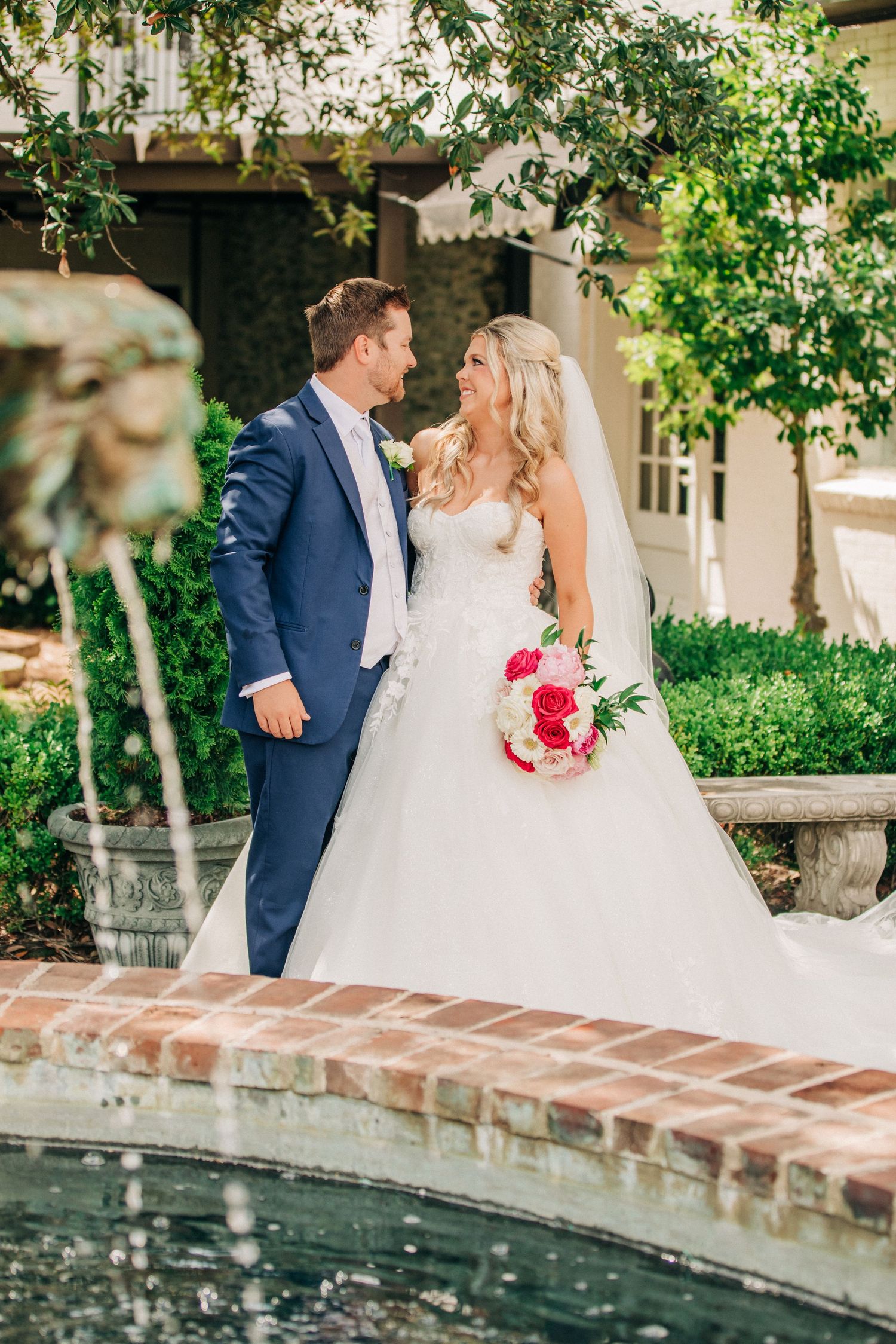 A bride in a white ballgown and groom in navy suit stand near a garden fountain on their wedding day.