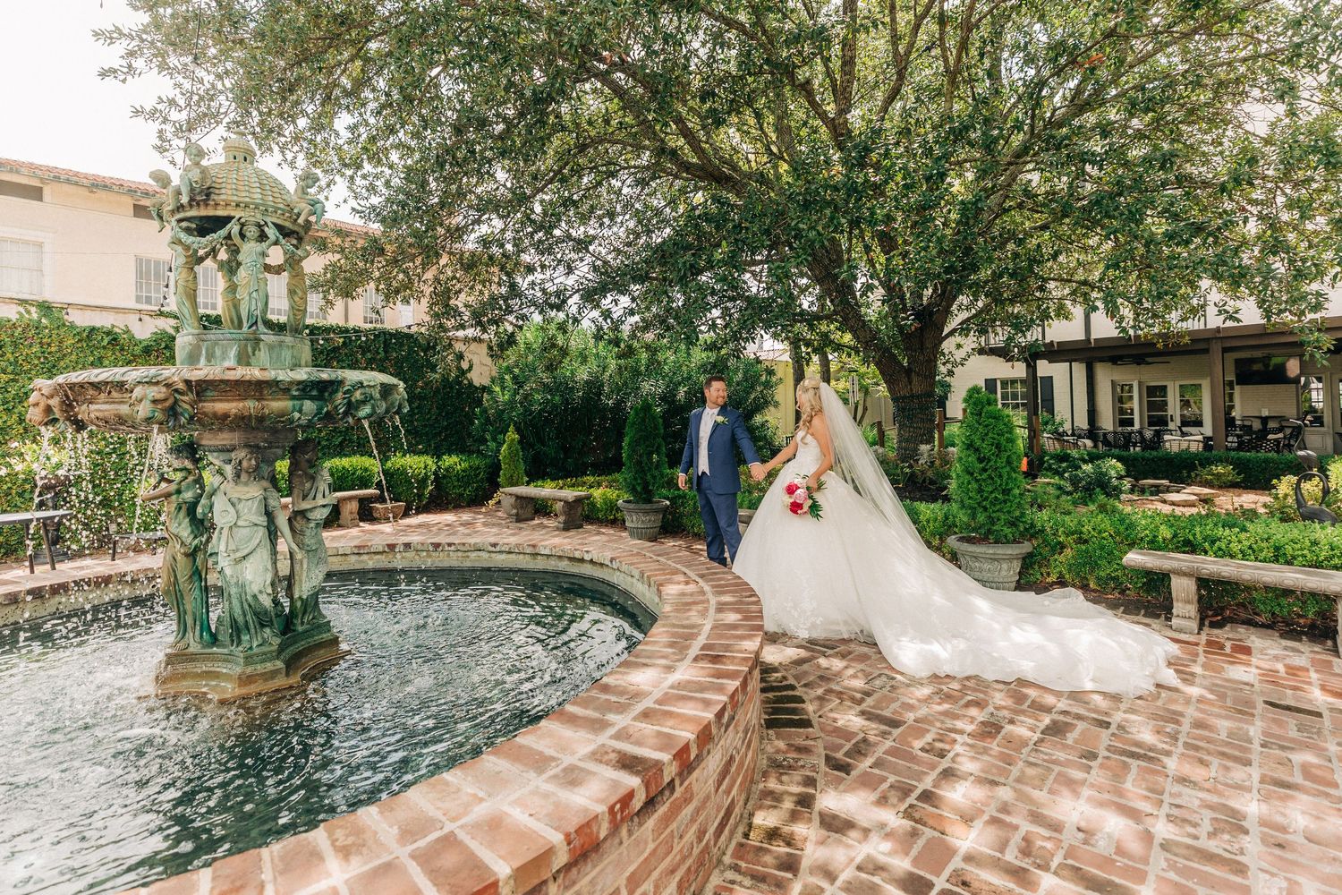A couple poses by an ornate stone fountain surrounded by lush gardens and brick pathways.