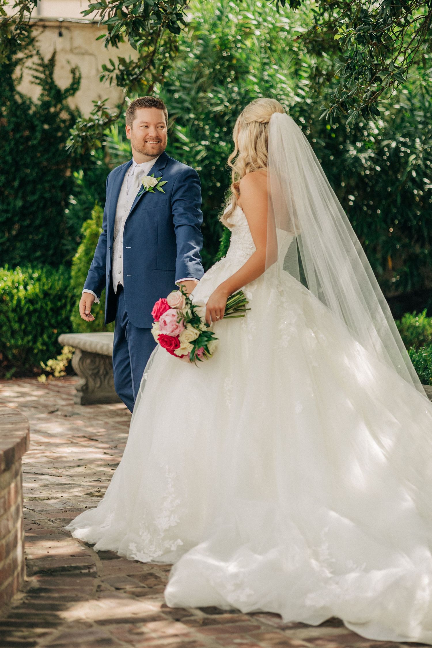 A wedding couple in navy suit and flowing white gown with pink bouquet pose in a lush garden setting.