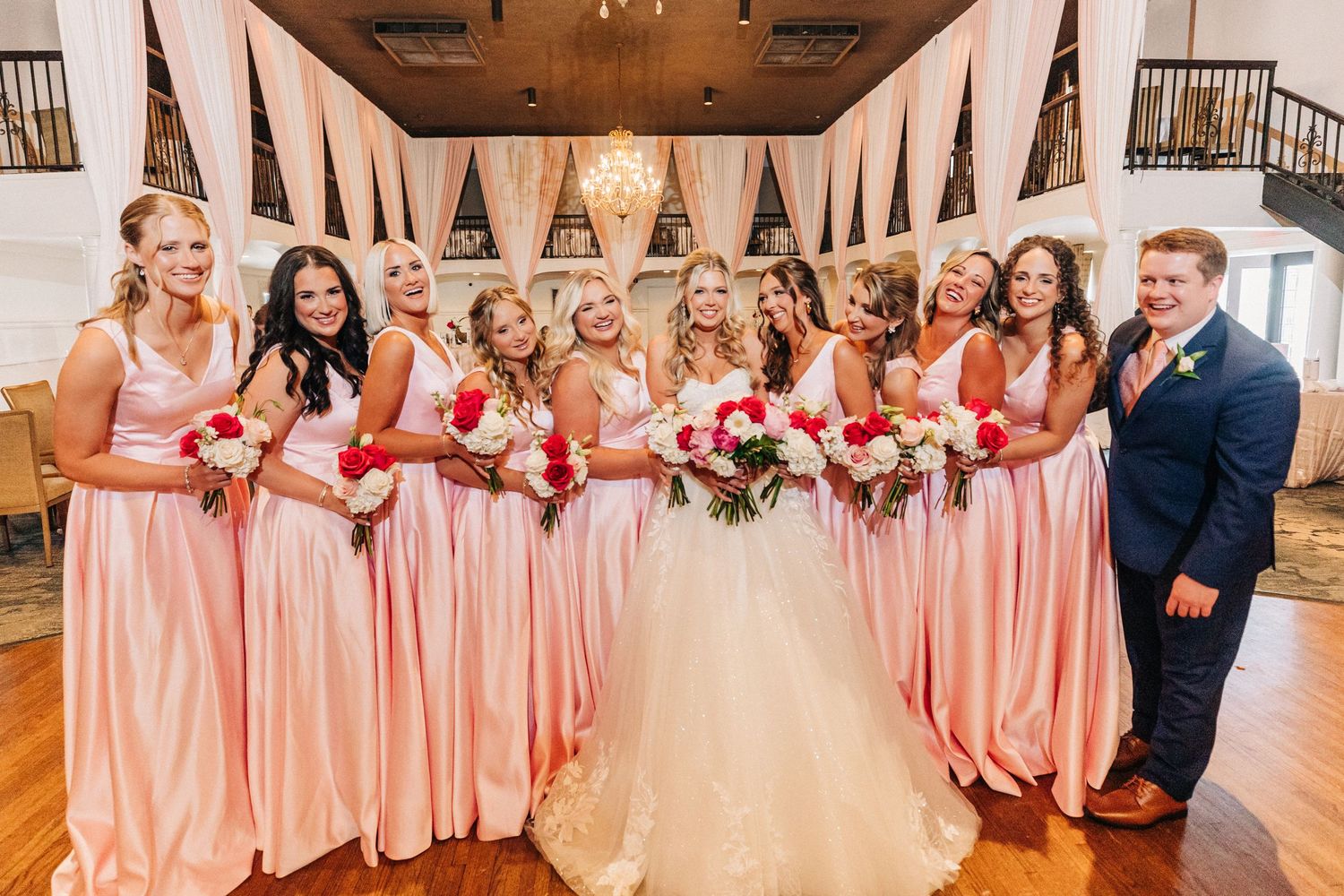 Wedding party wearing pink bridesmaid dresses gather together in an elegant wood-floored venue.