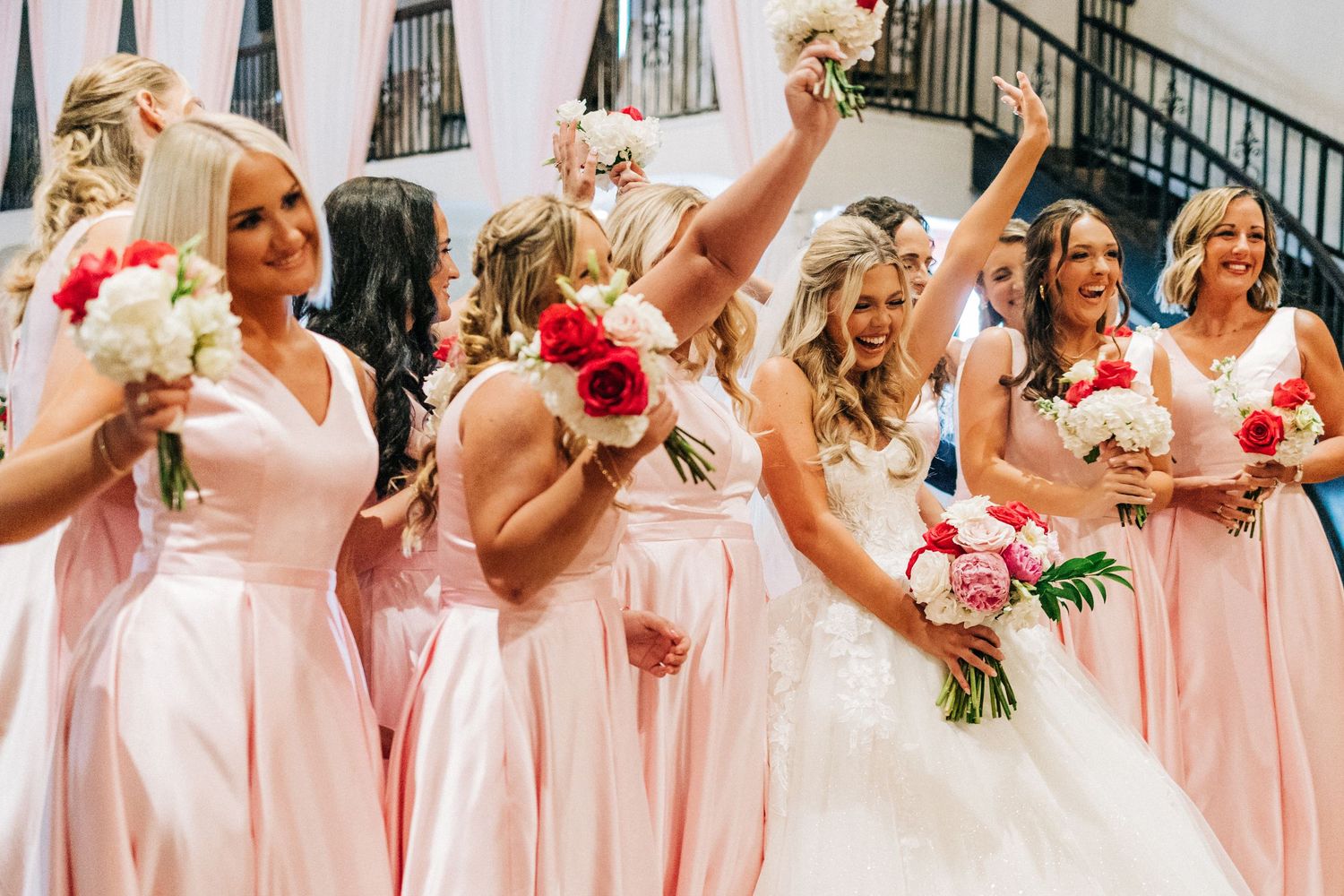 A row of bridesmaids in peach dresses holding pink and white floral bouquets smile and laugh during a wedding.