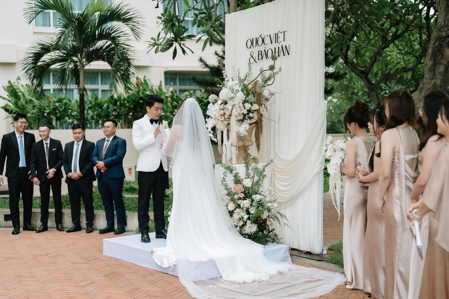 Outdoor wedding ceremony with groom reading vows at elegant white floral backdrop with groomsmen and bridesmaids.