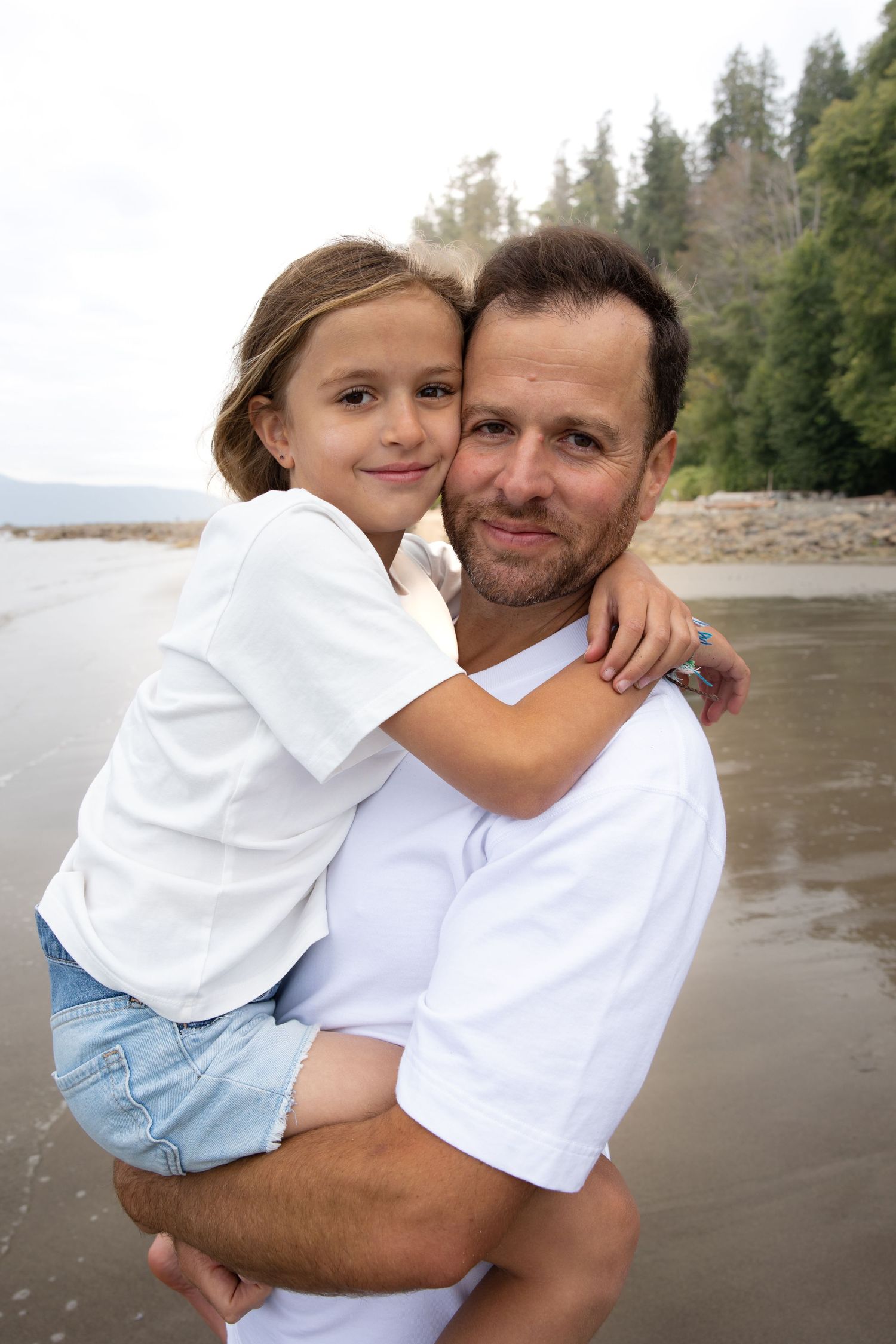 Parent and child in white shirts embrace while standing on a beach with evergreen forest in the background.