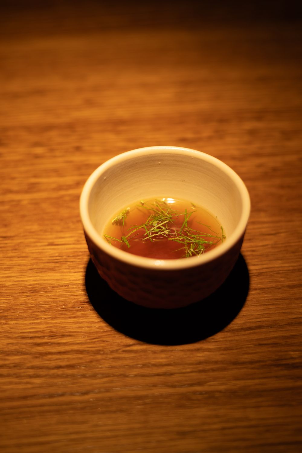 A small white ceramic bowl containing clear amber-colored broth with delicate herbs sits on a dark wooden table surface.