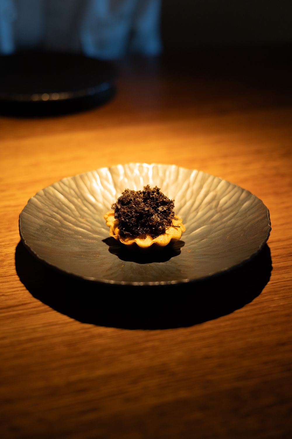 A small black caviar dish served on a textured white plate against a dark wooden surface with dramatic lighting.