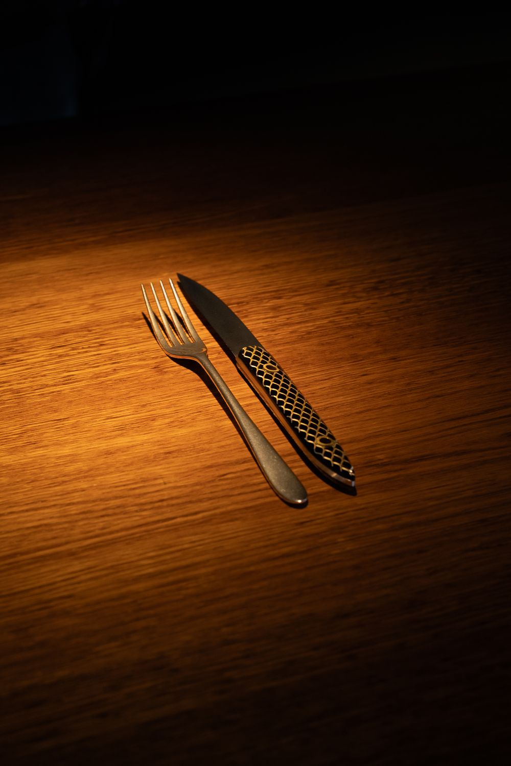 A knife and fork with decorative handles cast dramatic shadows on a warmly lit wooden table surface.