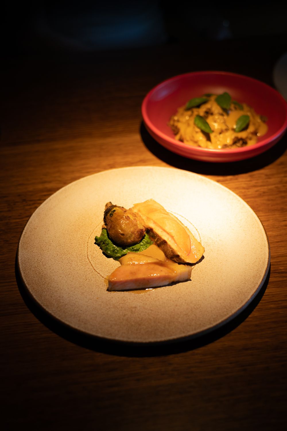 Elegant plated dish with seared meat on white ceramic plate with red bowl of garnish in dark lighting.