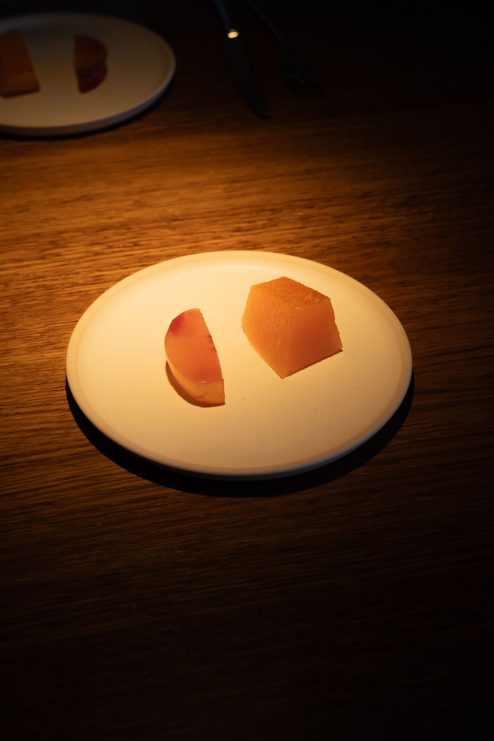 Two thin slices of prosciutto ham arranged on a white plate against a dark wooden table in dramatic lighting.