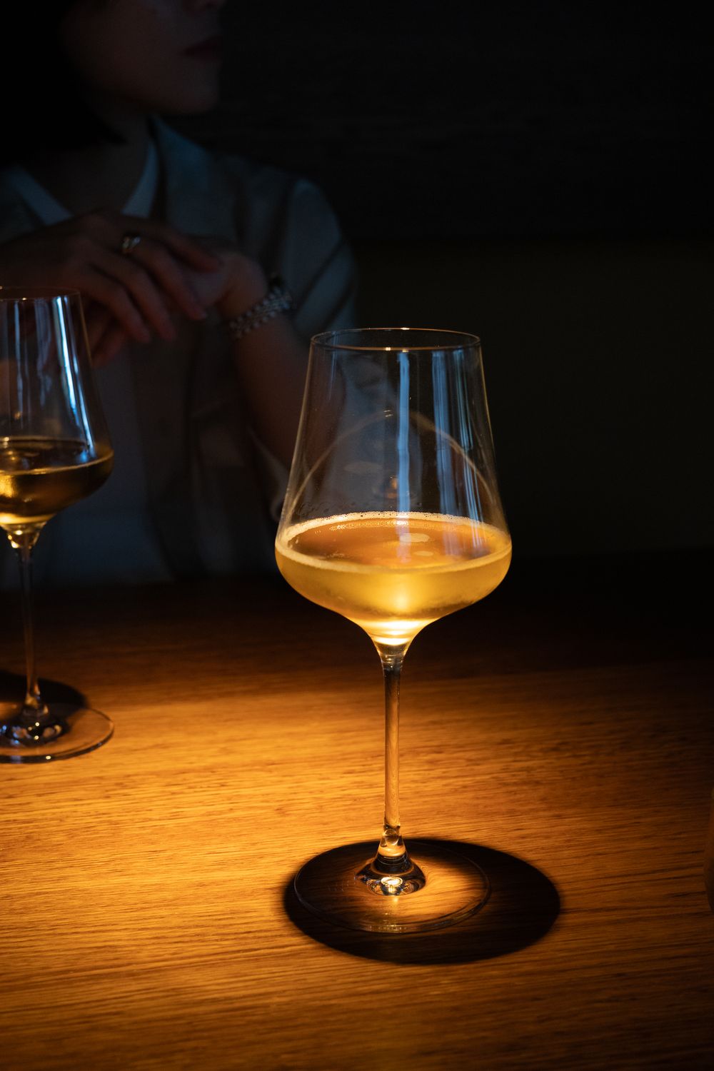 Wine glass casting dramatic shadows on wooden table in dim lighting with warm ambient glow.