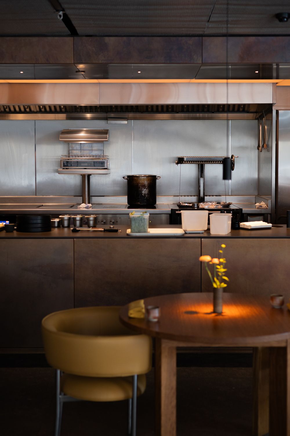 Modern restaurant interior with industrial kitchen setup, round wooden table, and yellow upholstered chairs in warm lighting.
