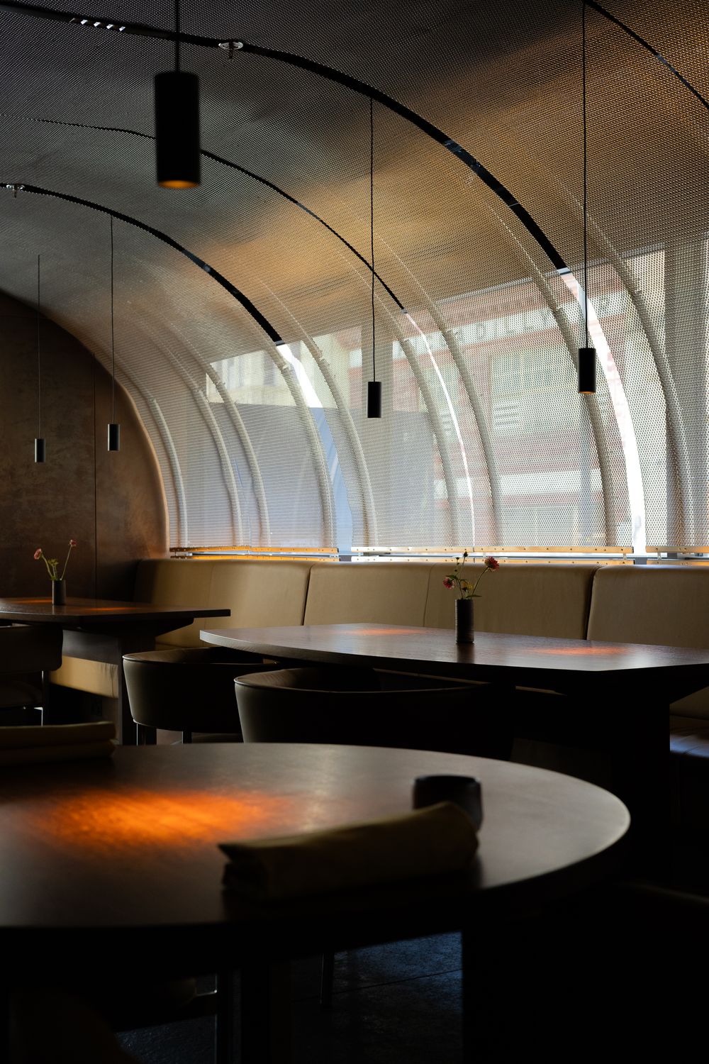 Modern restaurant interior with curved white walls, pendant lighting, and dark round tables in moody atmospheric lighting.