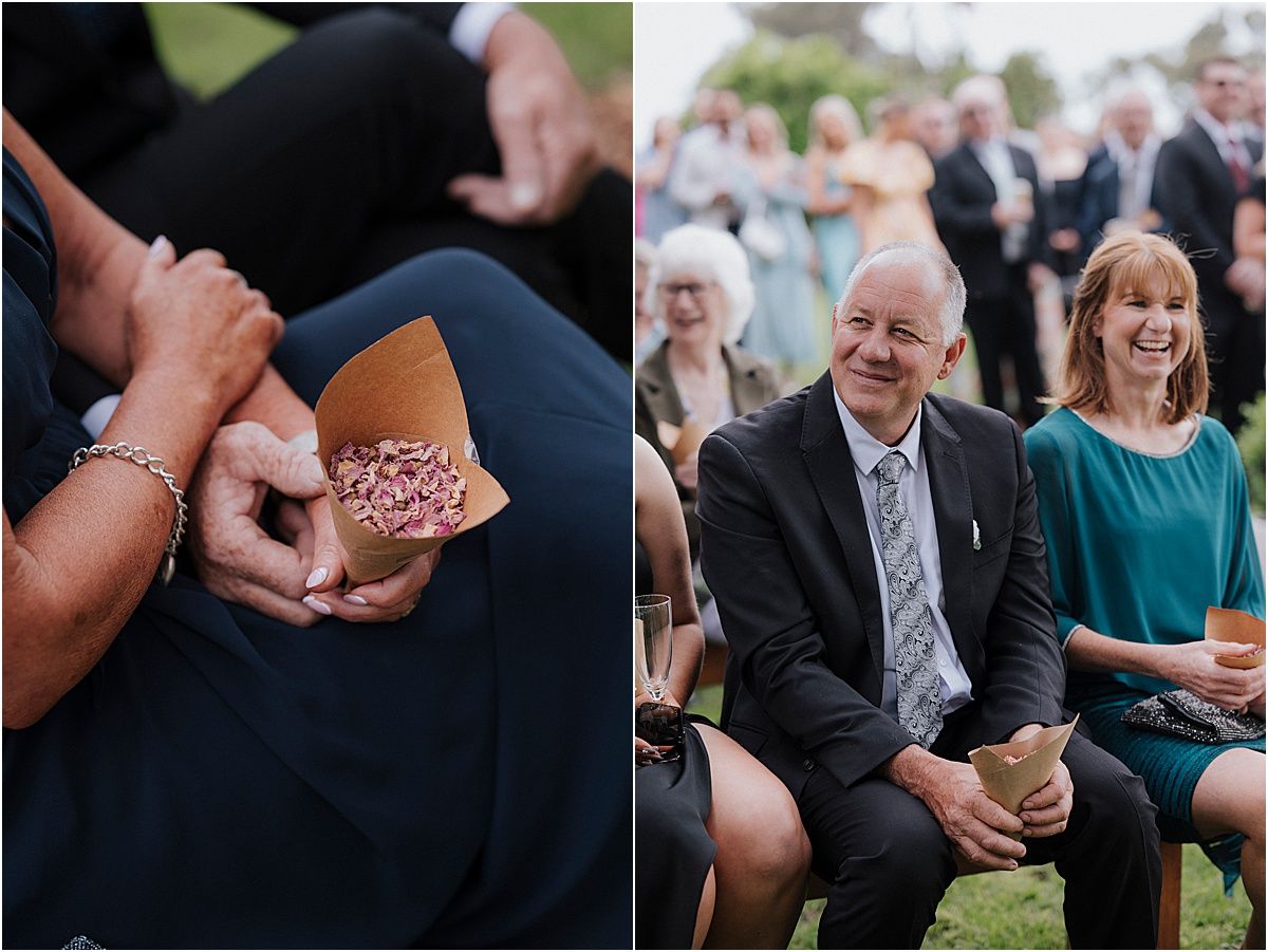 Wedding guests share an intimate moment looking at rings while others gather at outdoor ceremony.