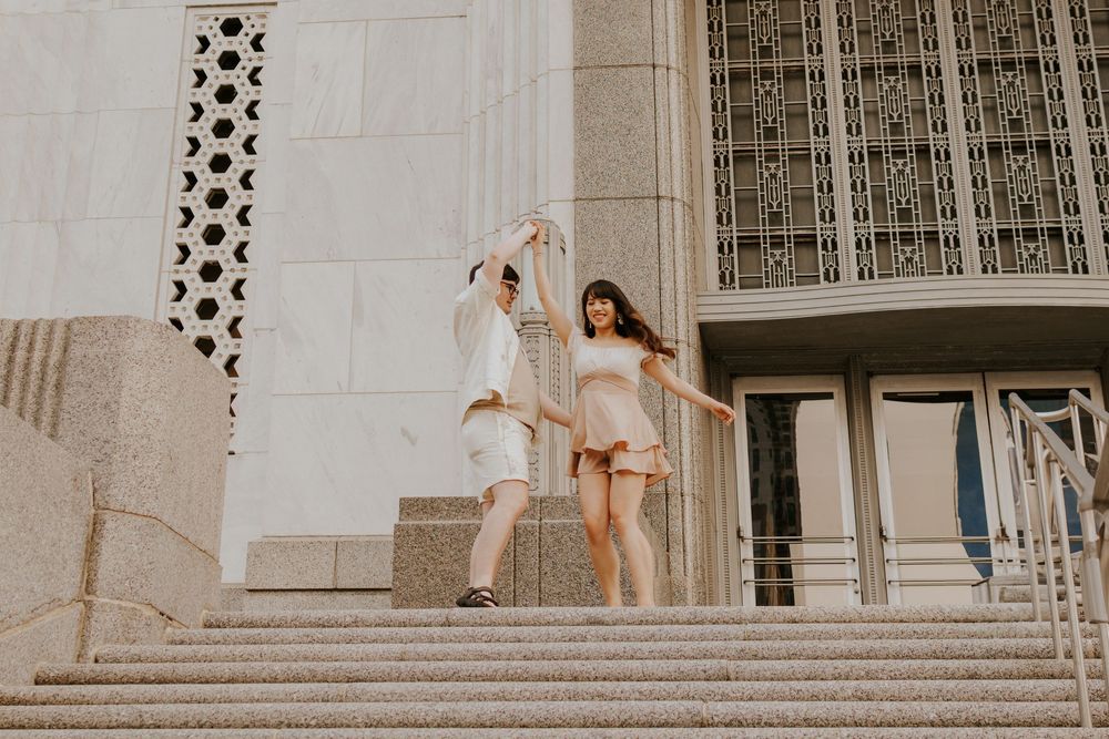 Two dancers in flowing outfits perform graceful movements on stone steps outside a building with architectural details.