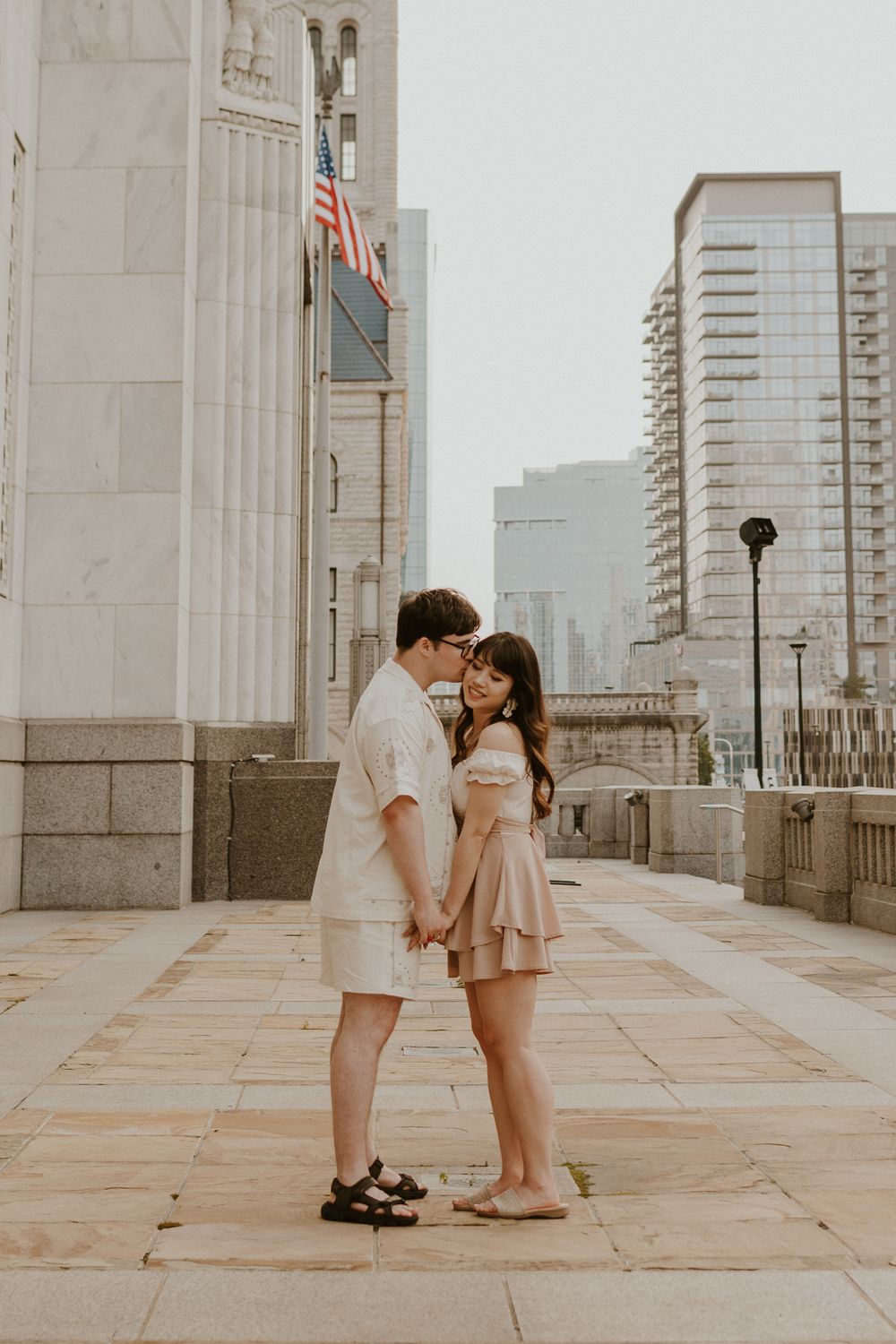 A couple shares an intimate moment against a cityscape backdrop on a pedestrian walkway with tall buildings visible.