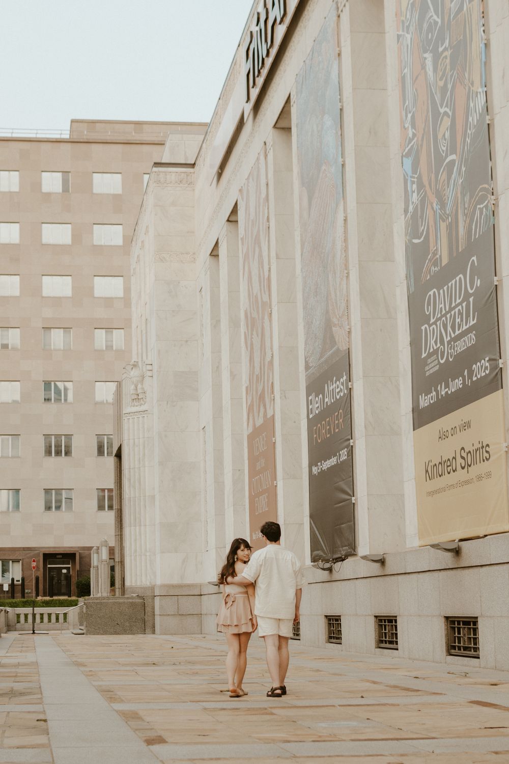 A couple in formal attire explores an architectural space with tall concrete pillars and modern design elements.