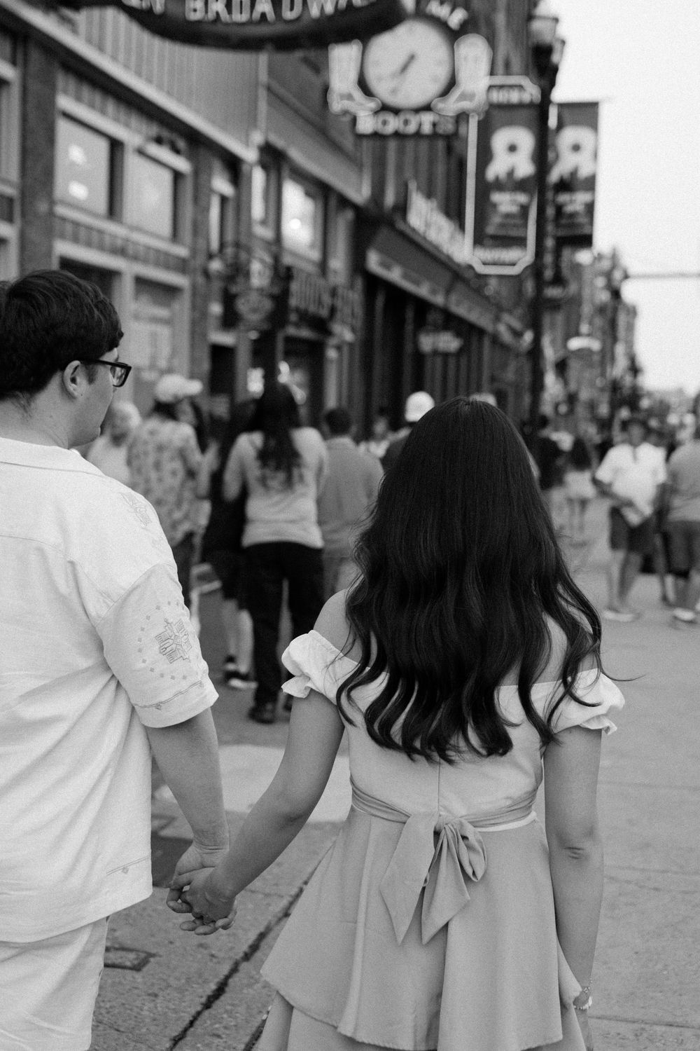 A couple walking together down a busy urban street with storefronts and pedestrians in black and white.