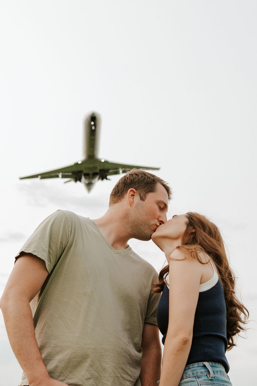 A romantic couple shares a kiss with a commercial airplane flying overhead against a white sky background.