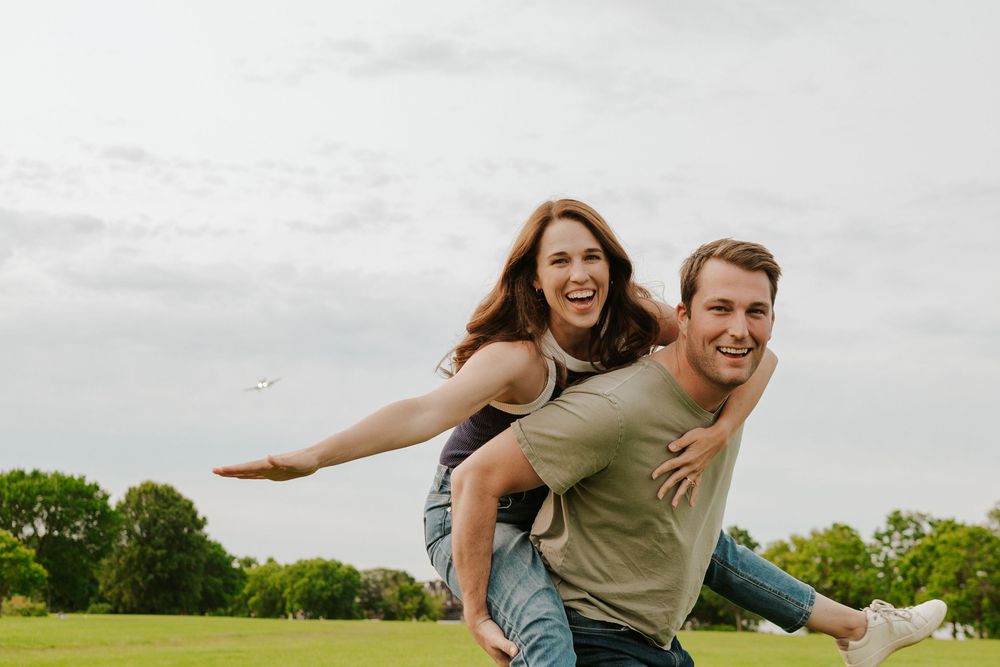 Two people enjoy a playful moment outdoors in a green field on a sunny day, giving piggyback rides and laughing.