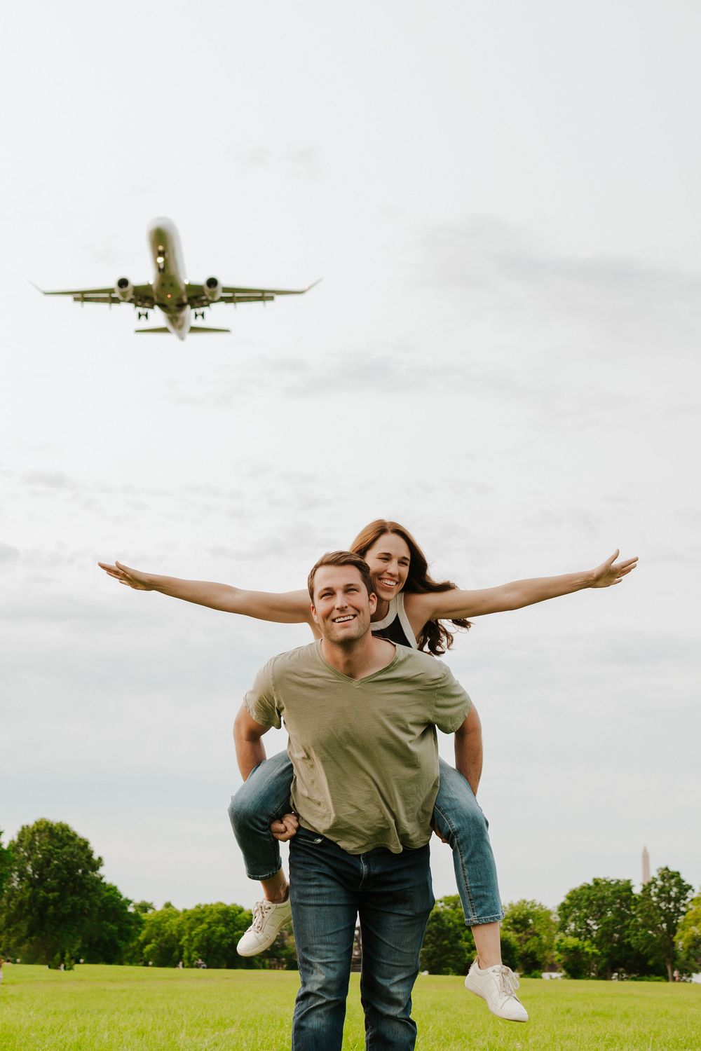 A person imitates airplane wings by riding piggyback on someone else as a plane flies overhead in a grassy field.