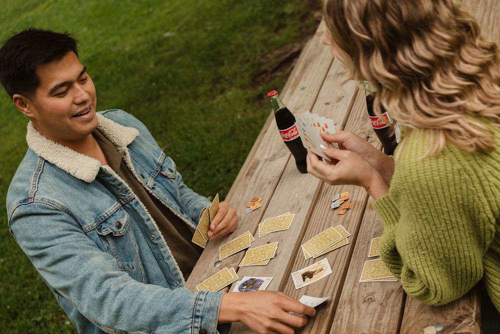 Two people in casual attire sit outdoors sharing beverages during a social gathering at a wooden picnic table.