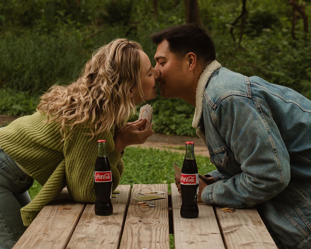 A couple shares a romantic moment over Coca-Cola bottles on a wooden picnic table in a lush green setting.