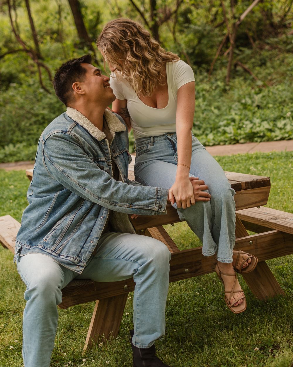 A couple shares intimate moments while sitting together on a wooden park bench surrounded by greenery.