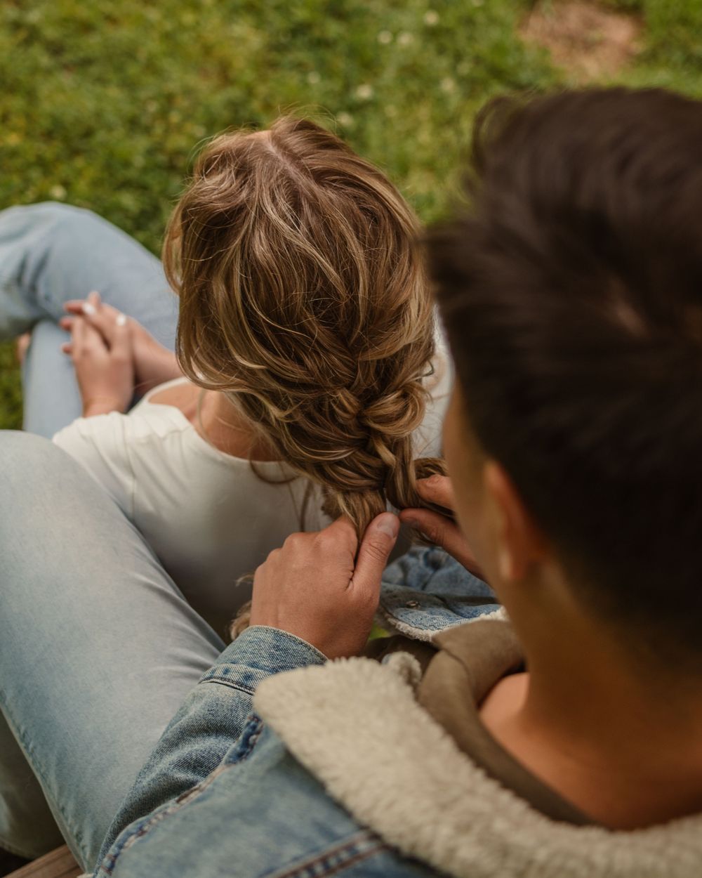Someone gently braiding hair while sitting together in casual denim and white clothing outdoors on grass.