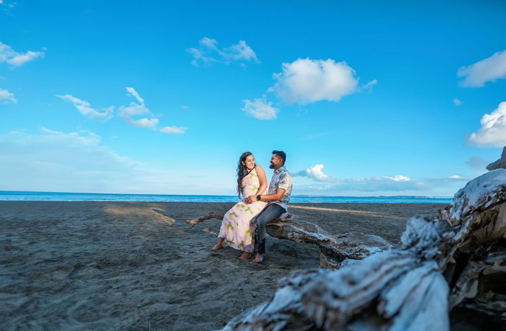 Two people sit together on dark volcanic rocks near the ocean under a bright blue sky and white puffy clouds.
