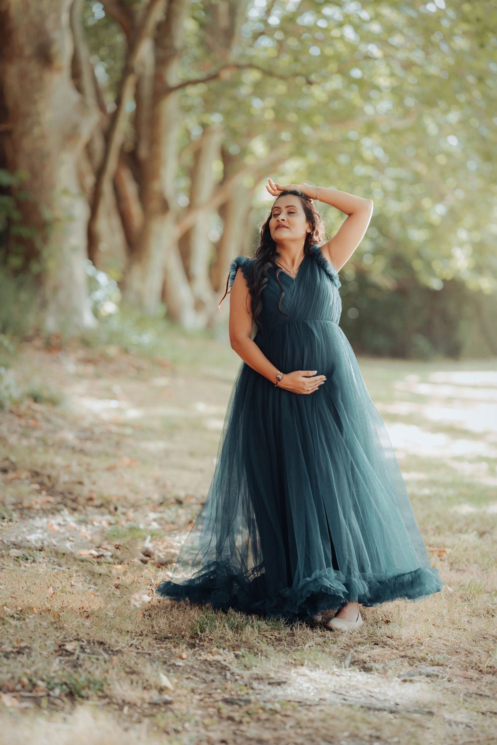 A figure in a flowing dark teal tulle gown poses gracefully against a dreamy outdoor woodland setting.