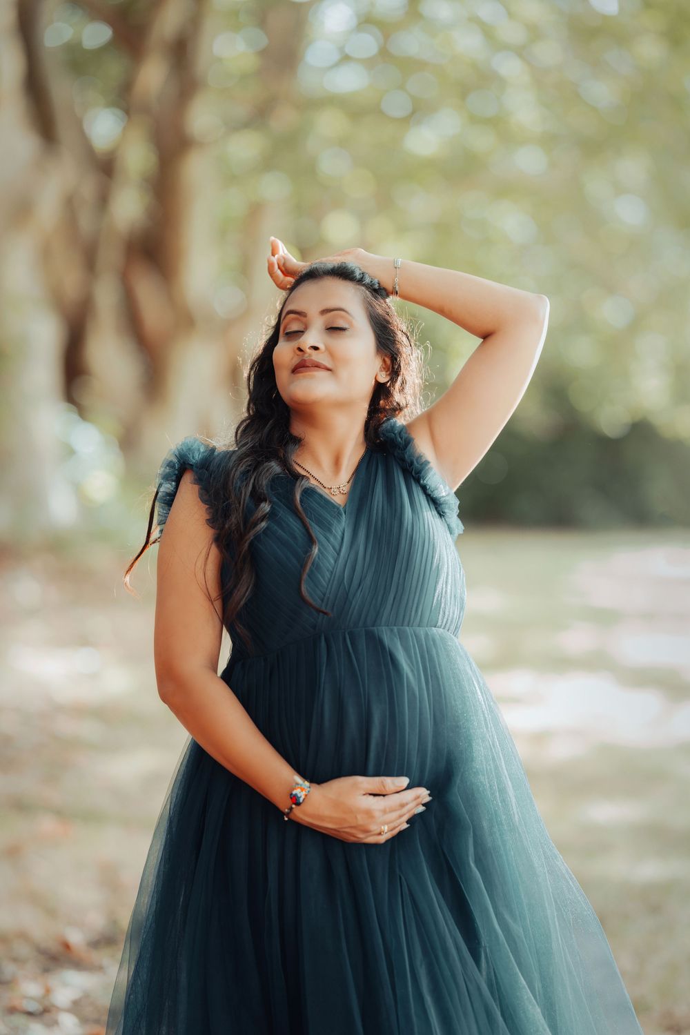 An expectant woman in a flowing teal tulle dress poses gracefully outdoors with soft bokeh lighting in the background.