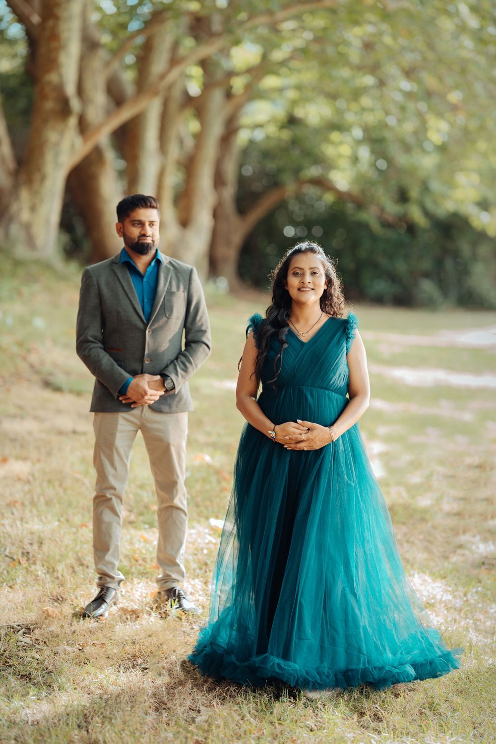 A couple in an outdoor photoshoot with a teal gown and grey blazer pose against trees in warm natural lighting.