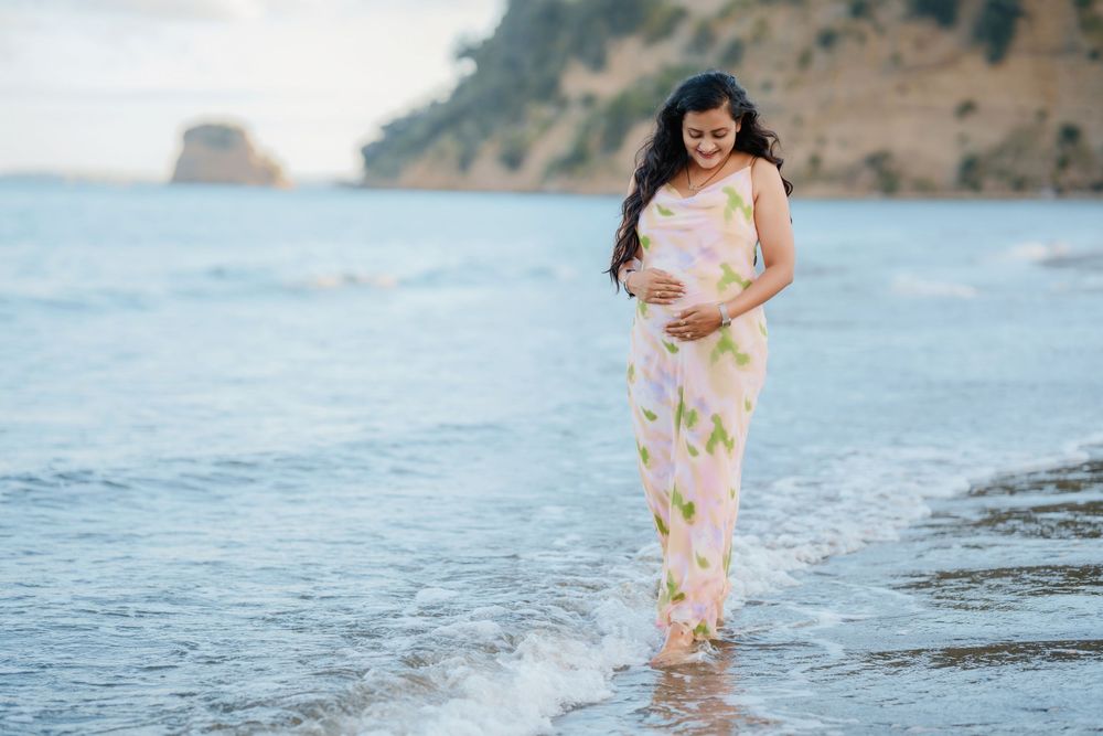 A figure in a pastel floral dress stands in gentle ocean waves at the beach with cliffs in the background.
