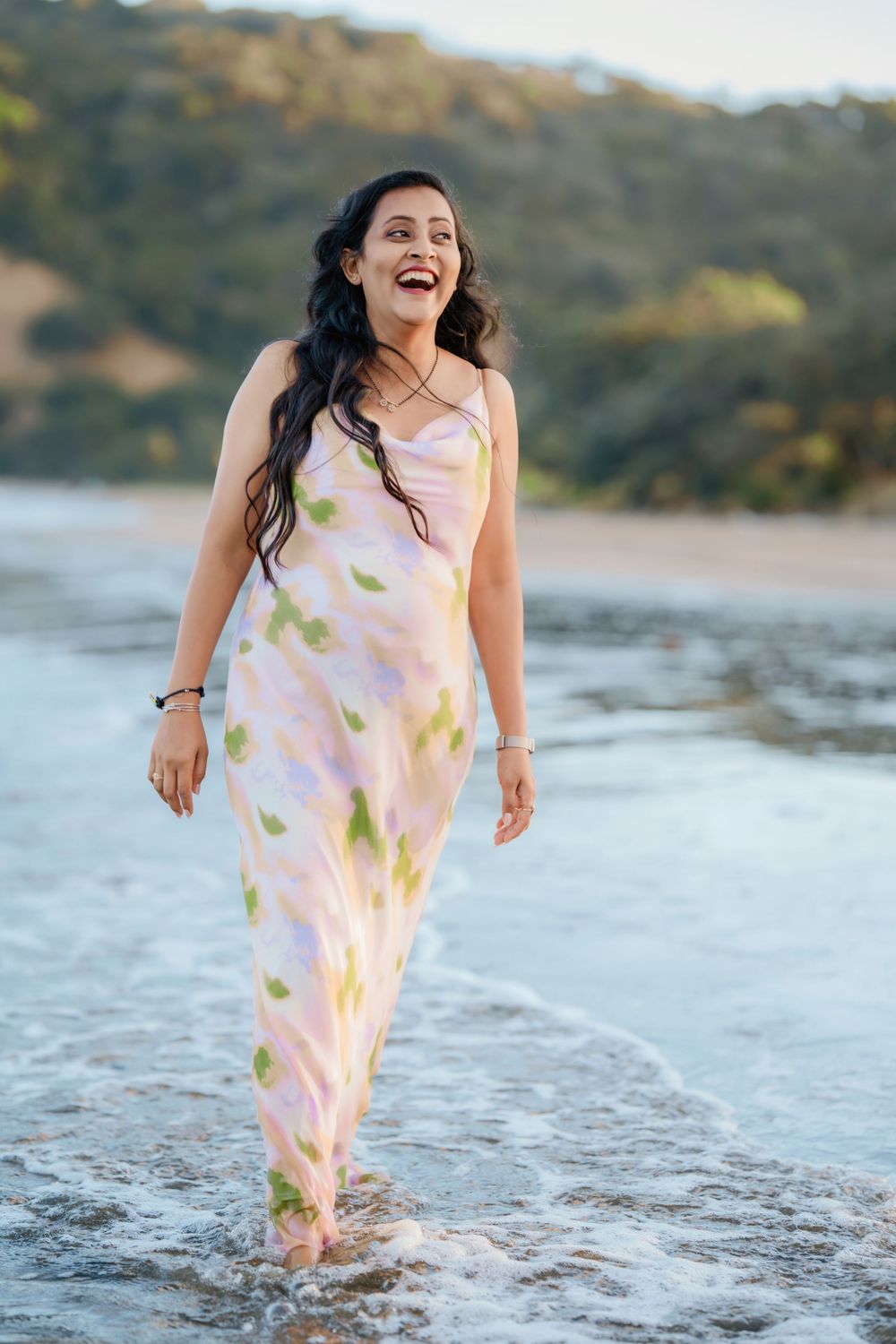 Someone wearing a flowy white and pink dress splashes joyfully through shallow ocean waves at sunset along the beach.