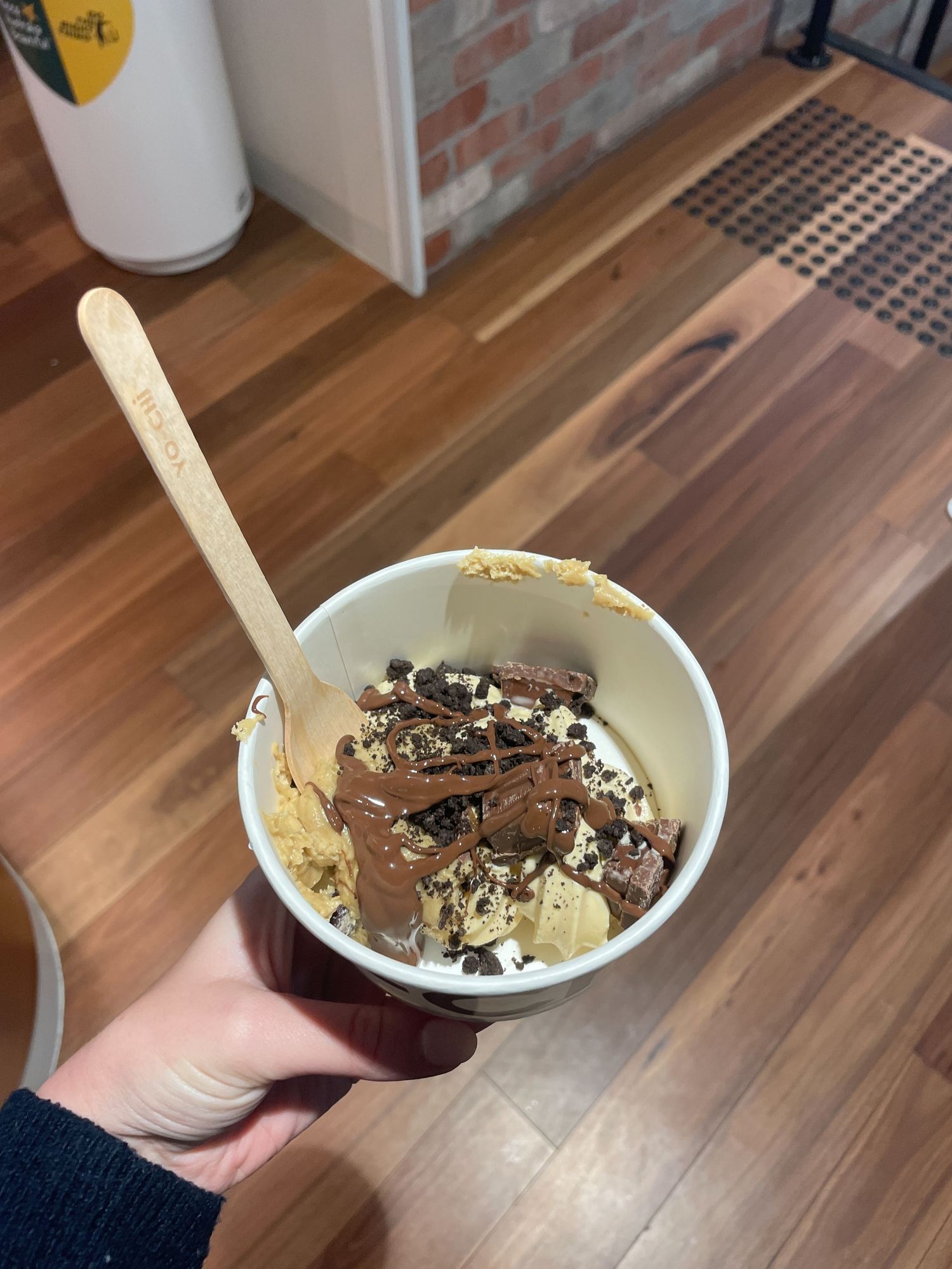 A bowl of chocolate ice cream topped with crushed cookies and nuts held over a wooden floor.