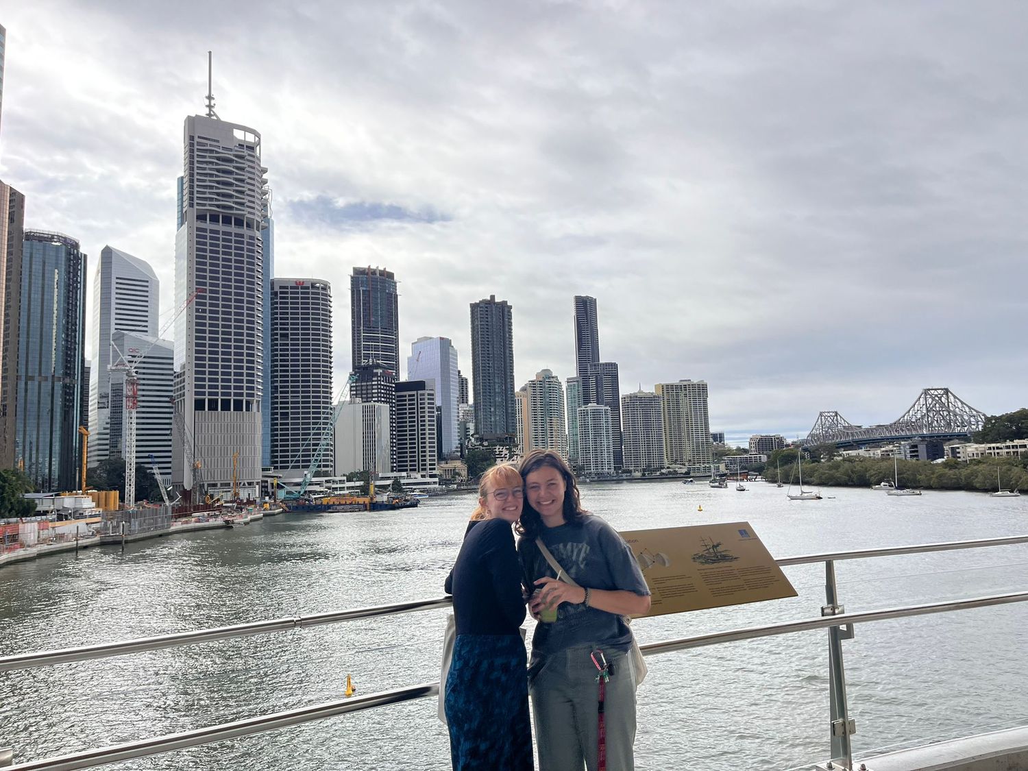 Two people stand along the Brisbane River with the city skyline and Story Bridge visible in the background.