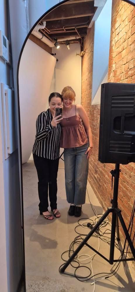 Two people in casual attire standing together in a narrow hallway with exposed brick walls and studio lighting equipment.