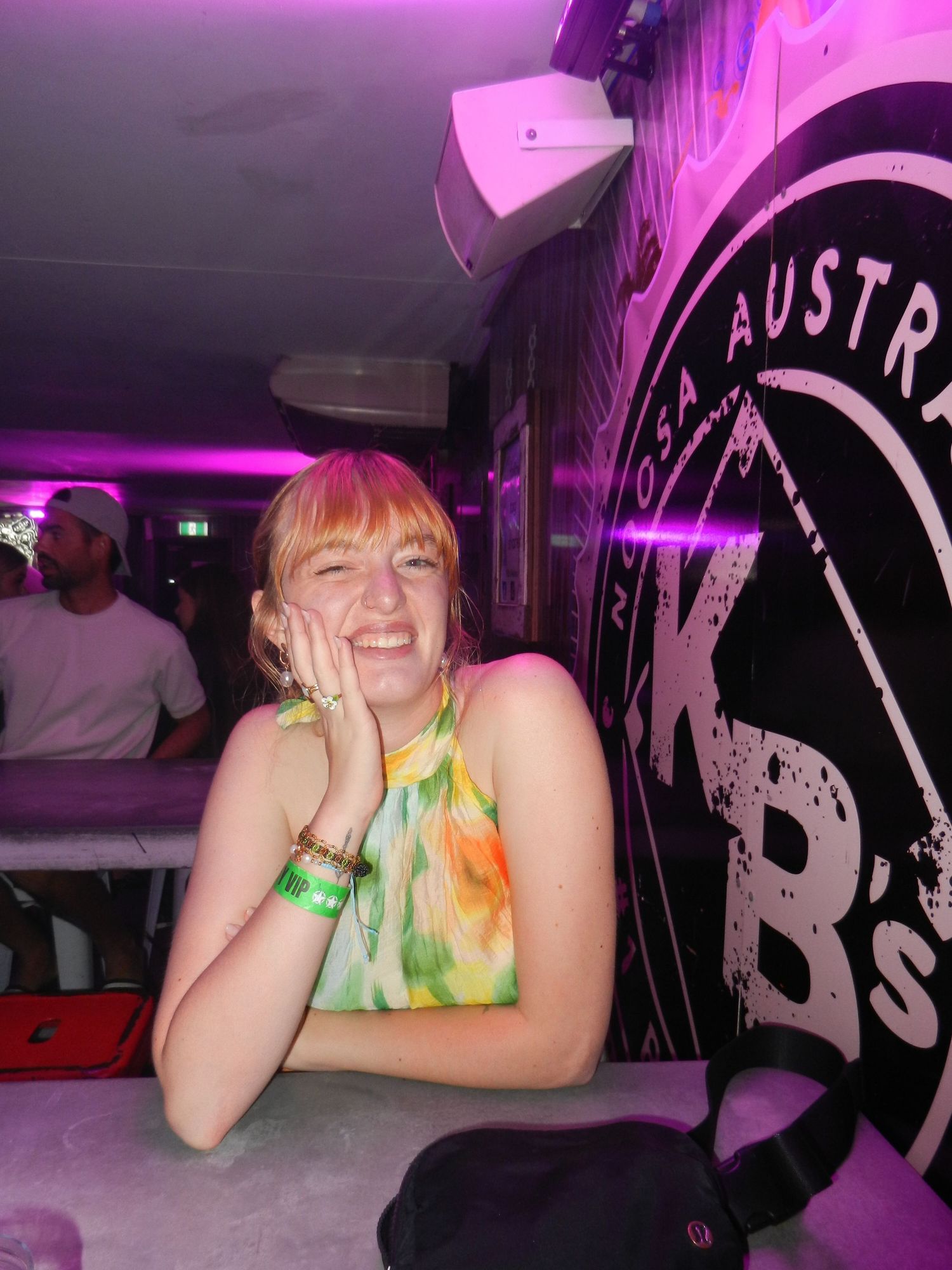 Person in colorful attire smiling at a nightclub with purple lighting and bar signage in background.