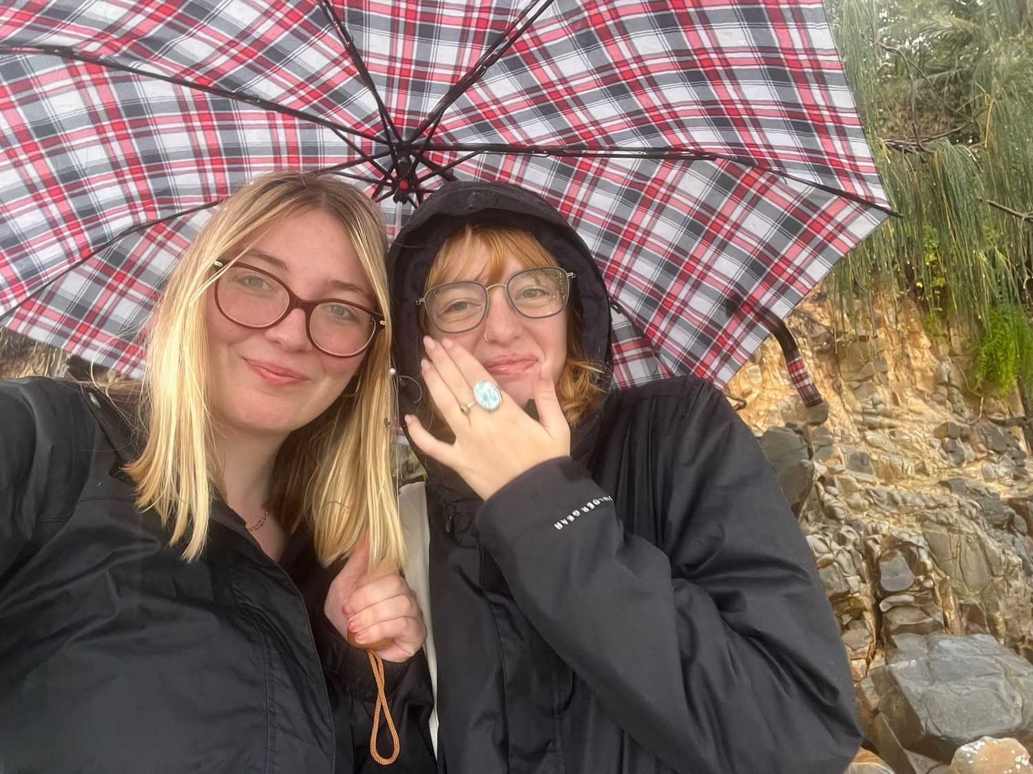 Two friends sharing a plaid umbrella on a cloudy day near rocky cliffs.