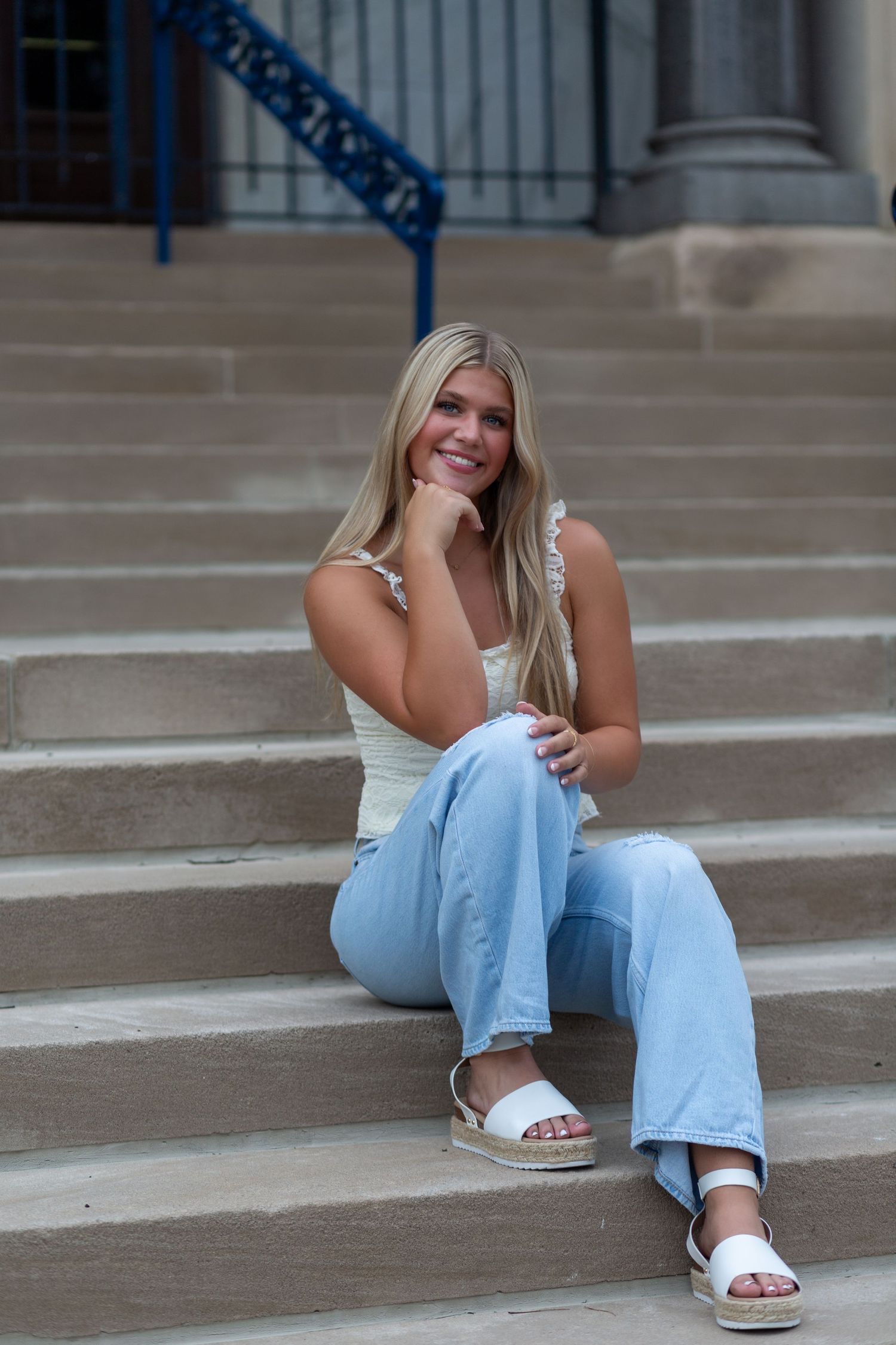 A person in a white tank top and light blue jeans sits casually on concrete steps while wearing white sandals.