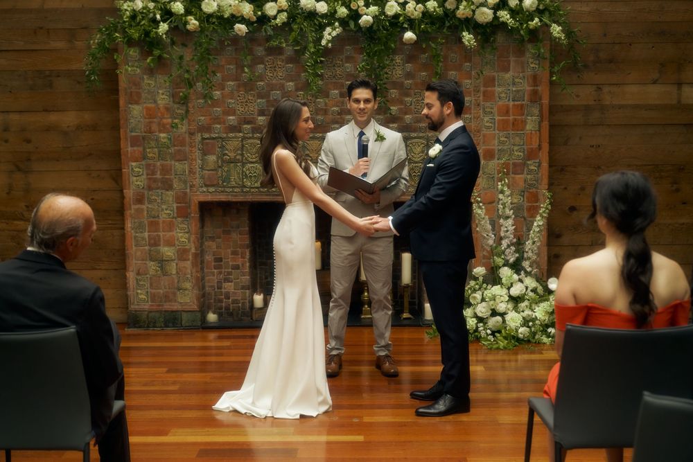 An intimate indoor wedding ceremony takes place in front of a rustic brick wall adorned with white floral arrangements at the Milling Room