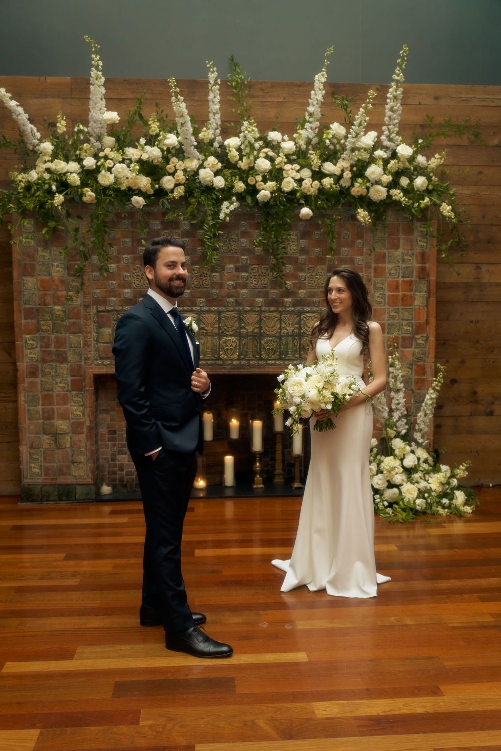 A couple stands in front of a brick fireplace decorated with elegant white floral arrangements and candles at the Milling Room