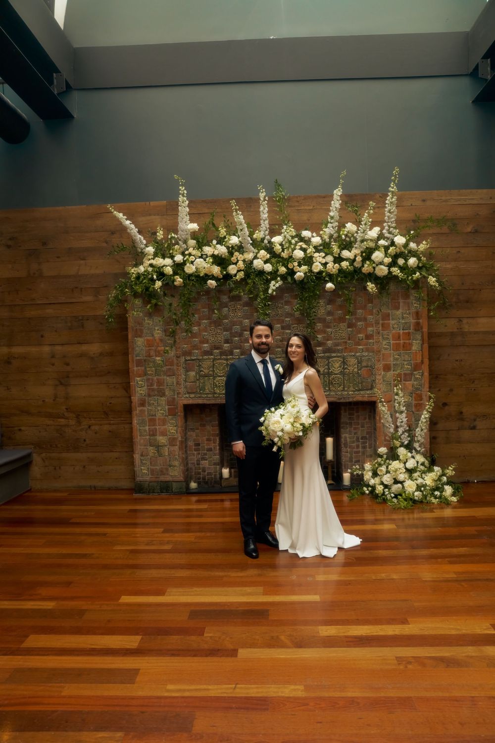 Elegant wedding ceremony setup with brick fireplace backdrop adorned with white floral arrangements and hardwood floors at the Milling Room