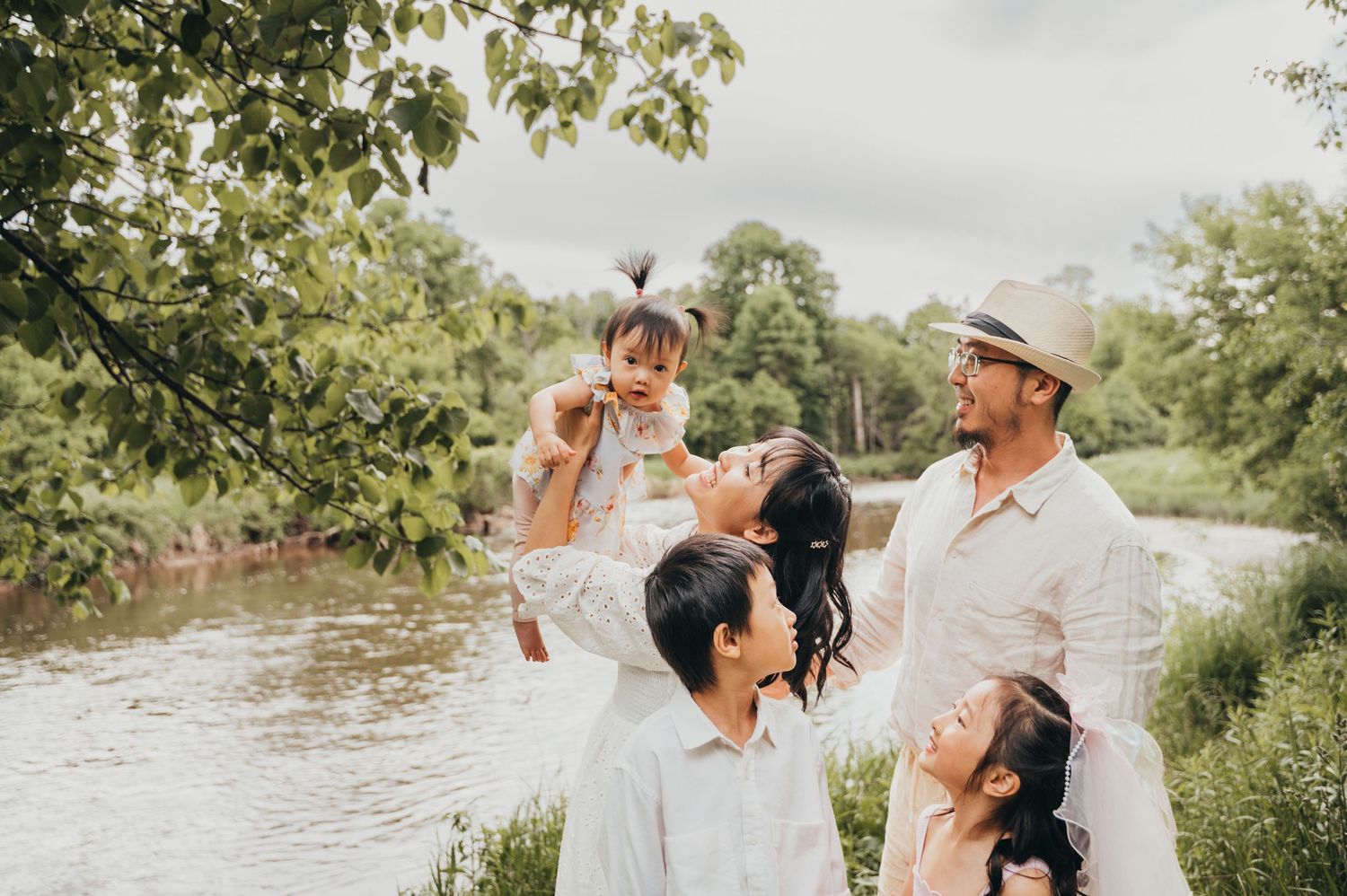 Mom holds baby up in air while Dad and kids look and smile in front of creek in Mississauga park