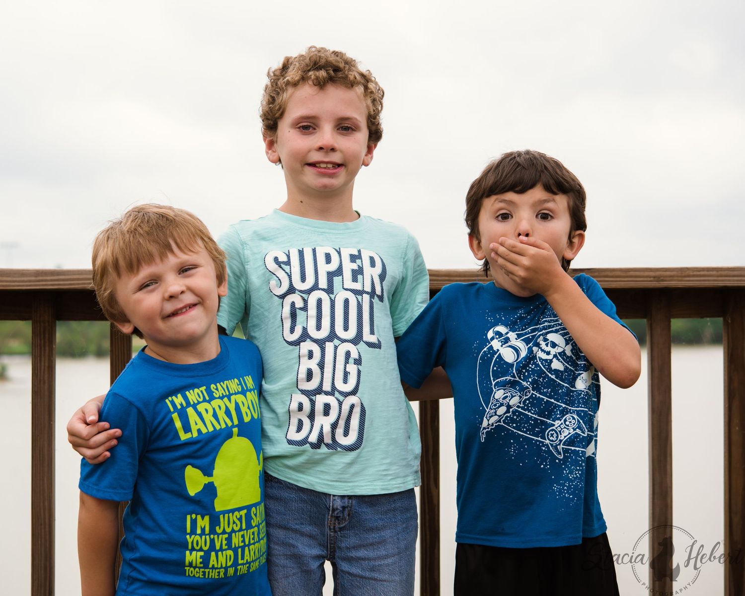 Three children in colorful t-shirts pose together on a wooden deck against an overcast sky.