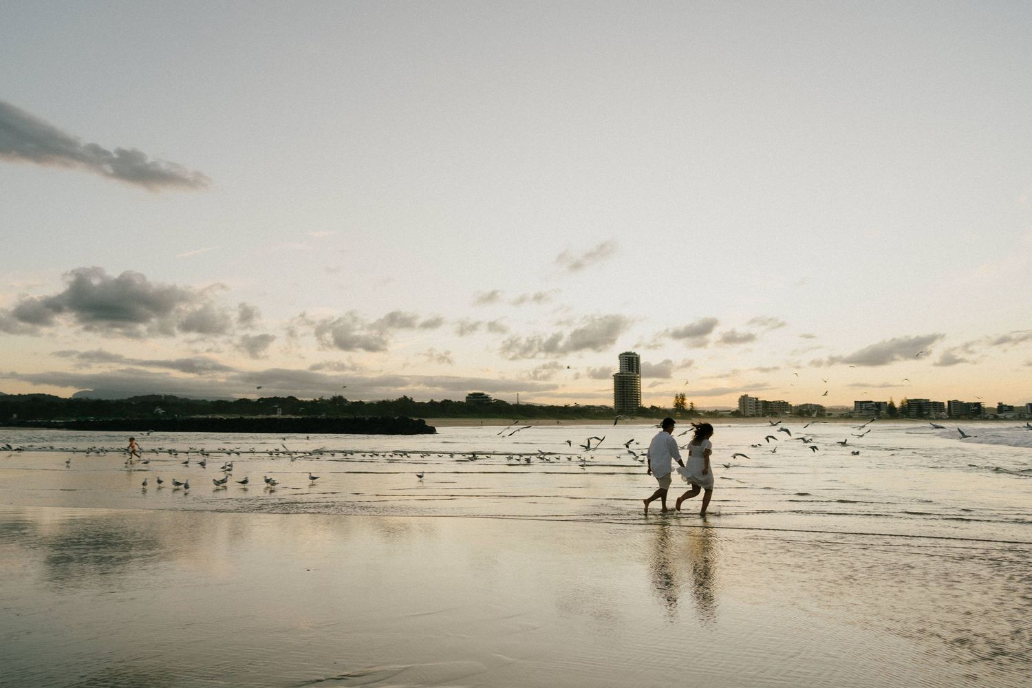 Silhouettes of people walking along a serene beach at sunset with reflective wet sand and dramatic cloud formations.