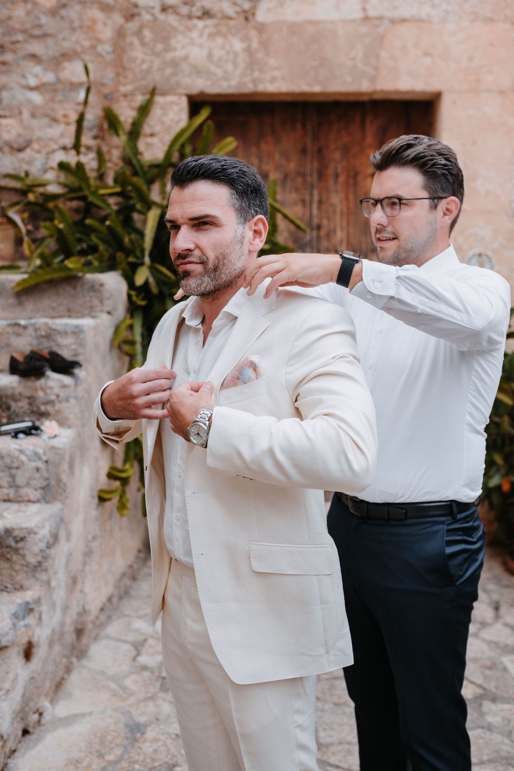Bräutigam und Trauzeuge bereiten sich auf die Hochzeit im Innenhof der Finca Xiclati vor, wobei einer dem anderen das Jackett anzieht.