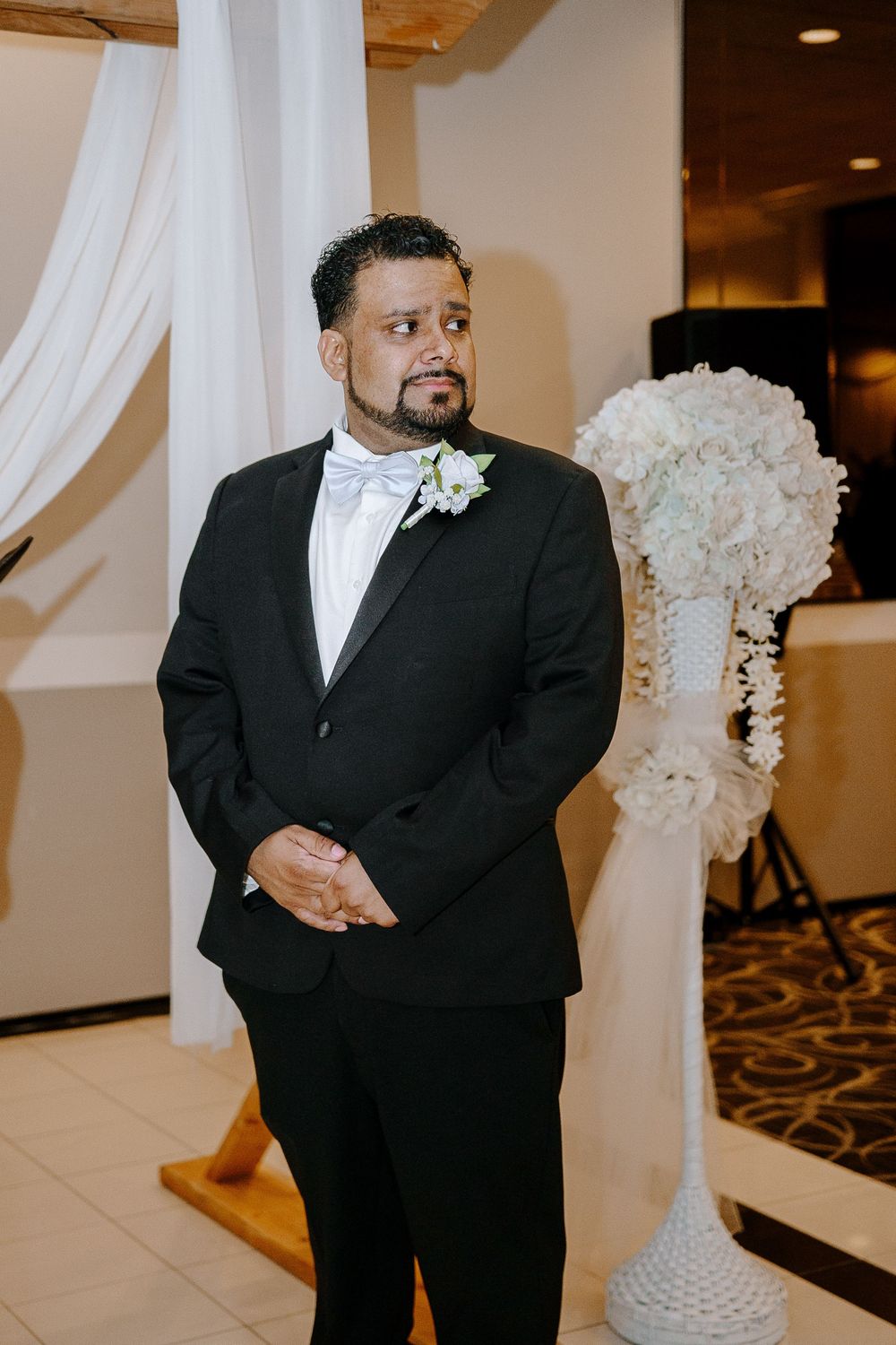 A groom wearing a black tuxedo and white bow tie smiles during a wedding ceremony.